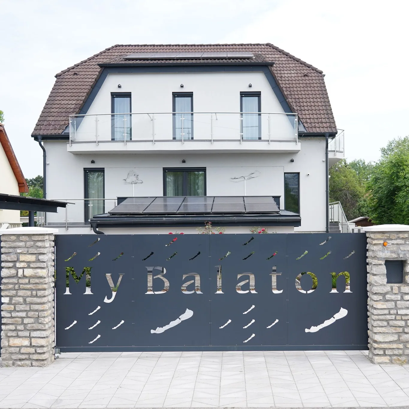A My Balaton háza a bejárati kapu felől, kivágott My Balaton felirattal, fehér homlokzattal és barna mansard tetővel